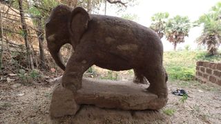 We see the stone-carved statue of an elephant with its trunk curved downward toward the ground. It is in a dirt-covered area of a grass field.