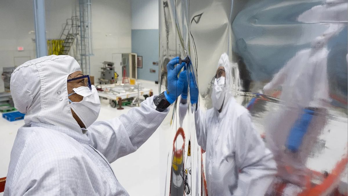 a technician in a white &#039;bunny suit&#039; examines a reflective silver metallic structure inside a large white-walled clean room