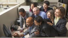 Office colleagues gather around one computer in a cubical to look at a computer screen. They are a diverse group and appear happy.