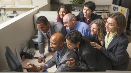 Office colleagues gather around one computer in a cubical to look at a computer screen. They are a diverse group and appear happy.