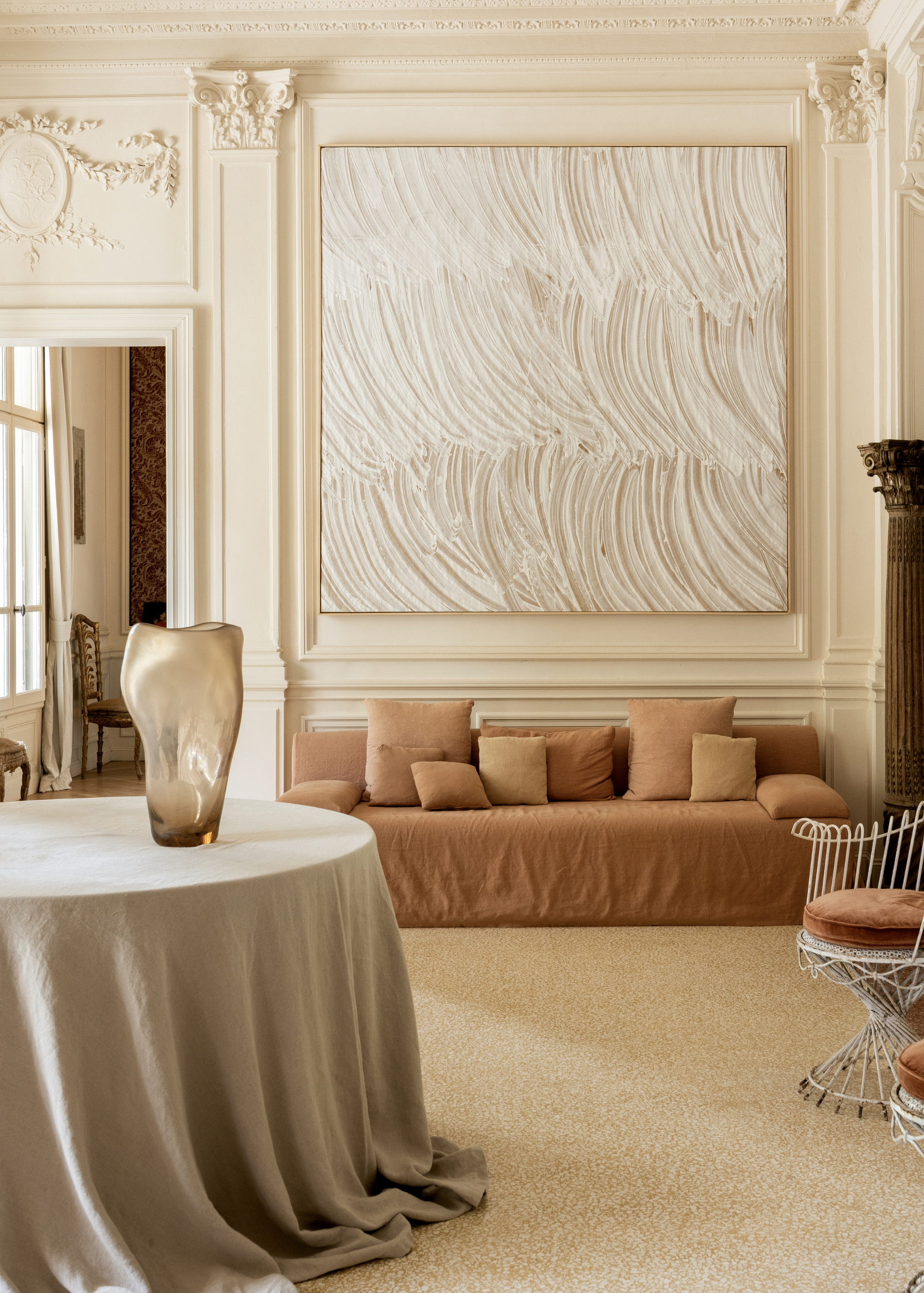 A living room with a textured painting, traditional trim, a brown sofa with many pillows, a round table draped in a cream tablecloth, and a pair of wire chairs