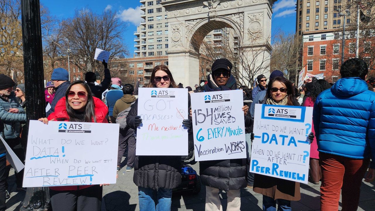 A group of people at the Stand Up For Science rally hold protest signs. One reads "Got measles? Me neither!". Another says "154 million lives saved every 6 minutes. Vaccination works".