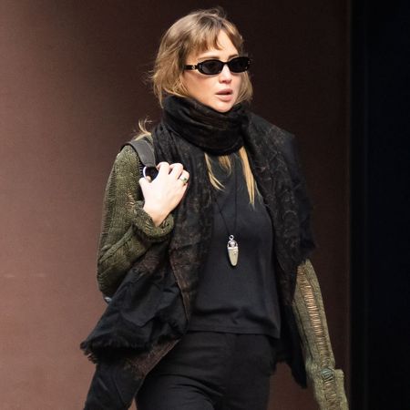 Jennifer Lawrence walks down the street in New York wearing black split-hem trousers with rust red satin shoes a black top, an oversized scarf and a green cardigan.