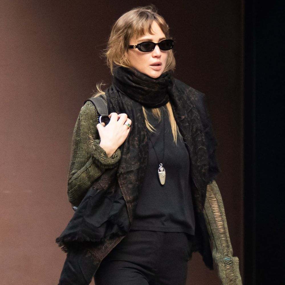Jennifer Lawrence walks down the street in New York wearing black split-hem trousers with rust red satin shoes a black top, an oversized scarf and a green cardigan.