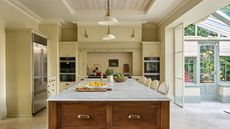 farmhouse style kitchen painted with butter yellow walls, a freestanding timber island with marble top, and a tiled area for the stove. There is an indoor conservatory next to the kitchen, panelled ceilings, and pendant lights hanging over the island