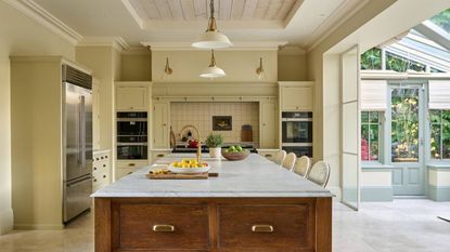 farmhouse style kitchen painted with butter yellow walls, a freestanding timber island with marble top, and a tiled area for the stove. There is an indoor conservatory next to the kitchen, panelled ceilings, and pendant lights hanging over the island 