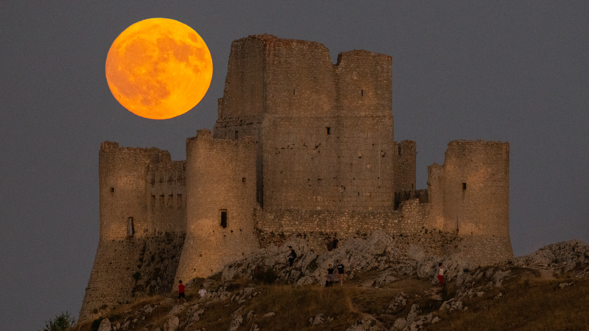 An orange full moon is pictured above the ruins of a castle parched on a rocky hill top.
