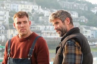 Sam Morgan (Louis Martin) and Adam Morgan (Dan Skinner) stand on the harbour in Shipton Abbott, with Sam facing the camera and Billy facing to the left. Sam is looking at Billy out the corner of his eyes, and there is clear tension between them.