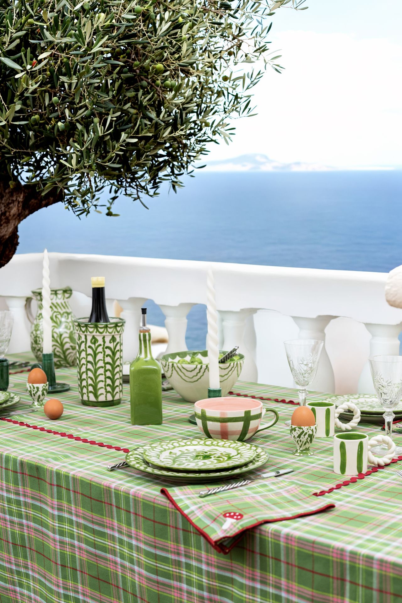 Image of a table on a white balcony that faces the sea. The table has a pink and green check tablecloth on it and is set with green and white ceramic tableware.