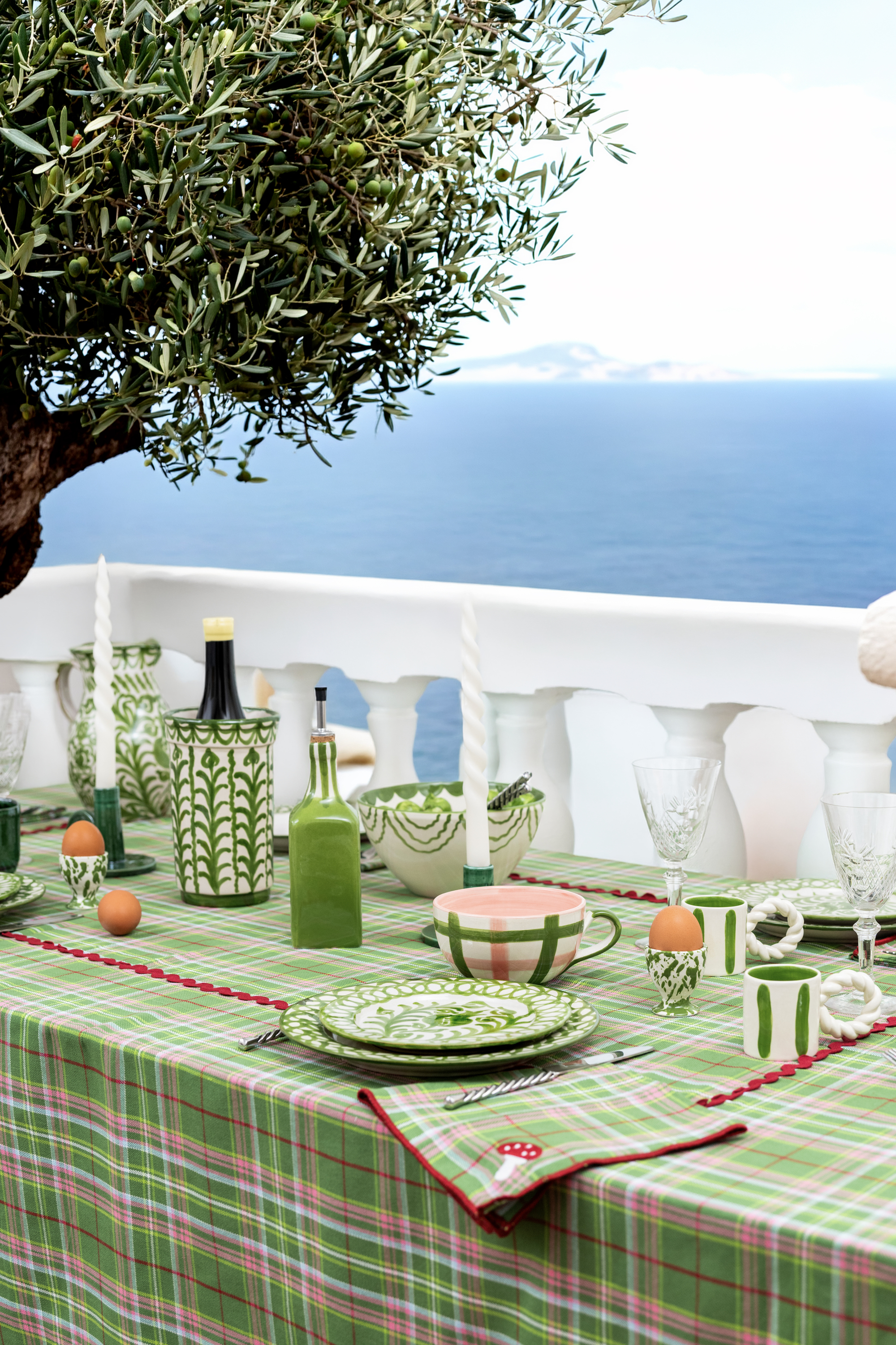 Image of a table on a white balcony that faces the sea. The table has a pink and green check tablecloth on it and is set with green and white ceramic tableware.