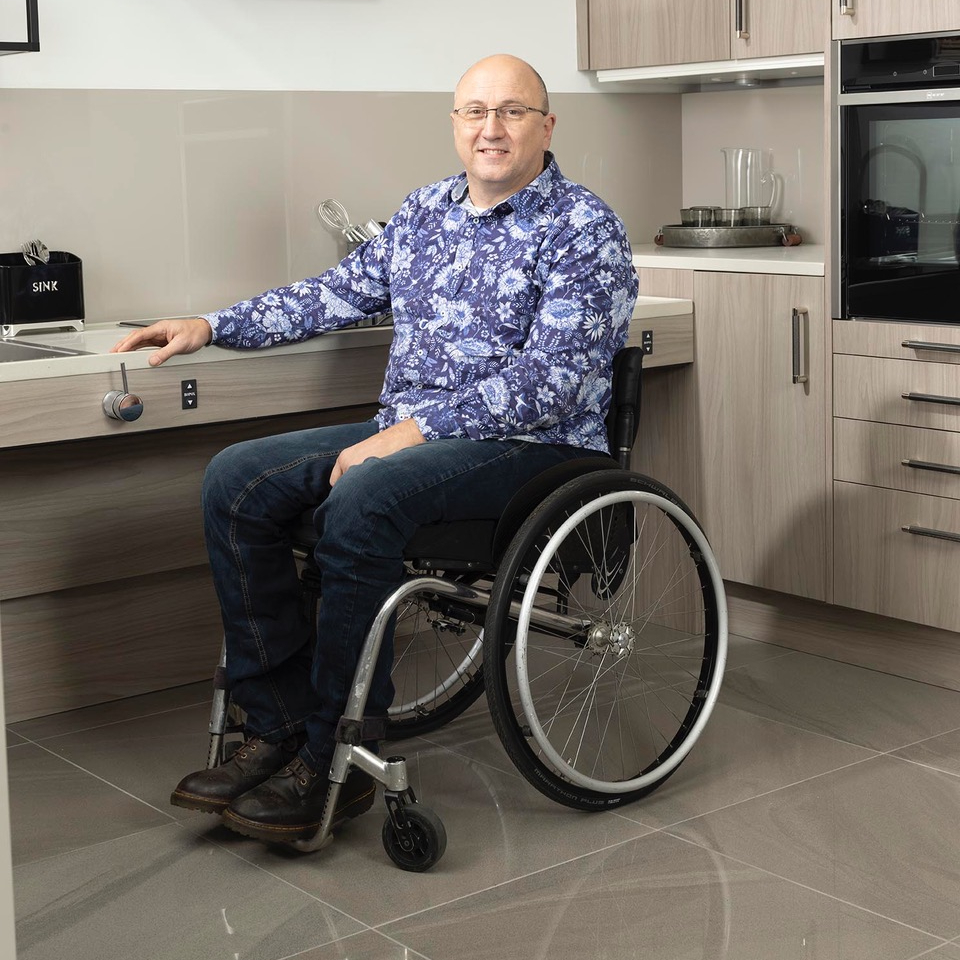 male wearing blue floral shirt and jeans sat in wheelchair in accessible kitchen