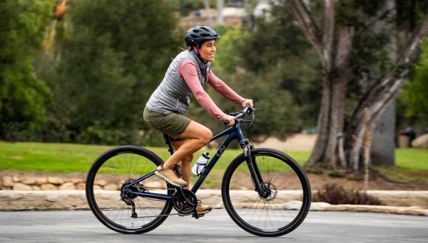  woman of colour cycles a hybrid bike, smiling, along the road. She&#039;s wearing shorts, a gilet and a helmet.