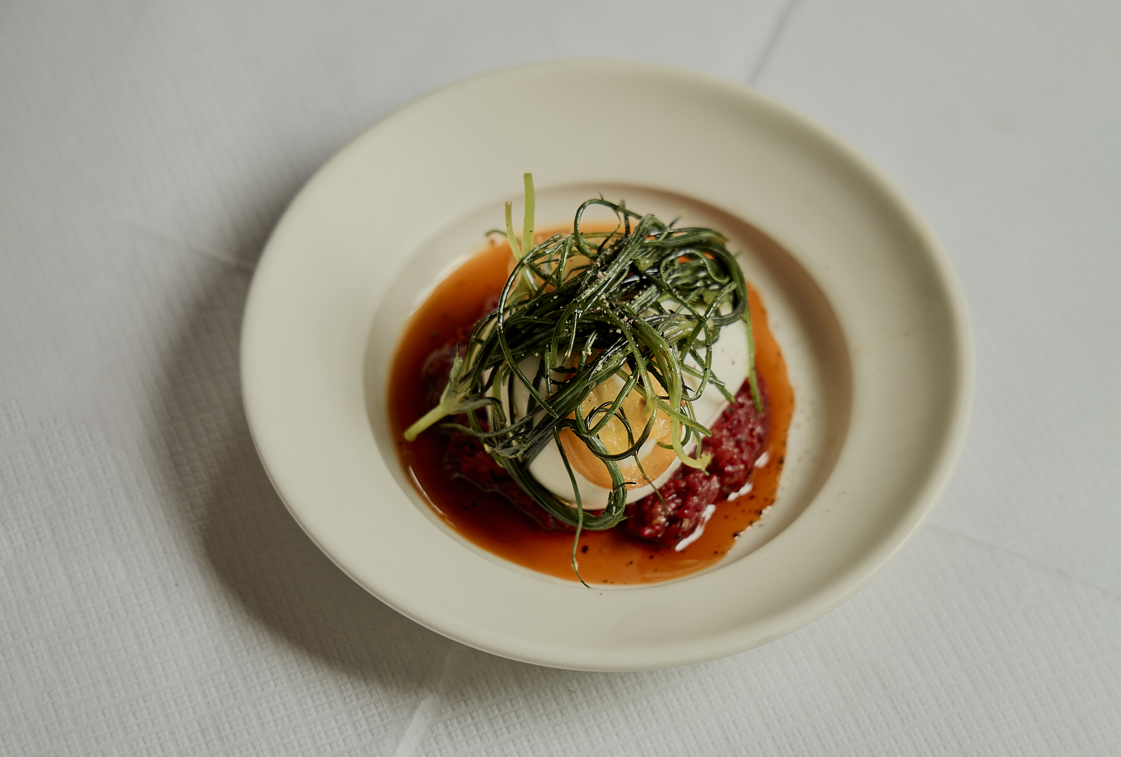 A vegetarian dish of fermented peppers and greens placed on top of a burrata in a cream-tinted ceramic dish.