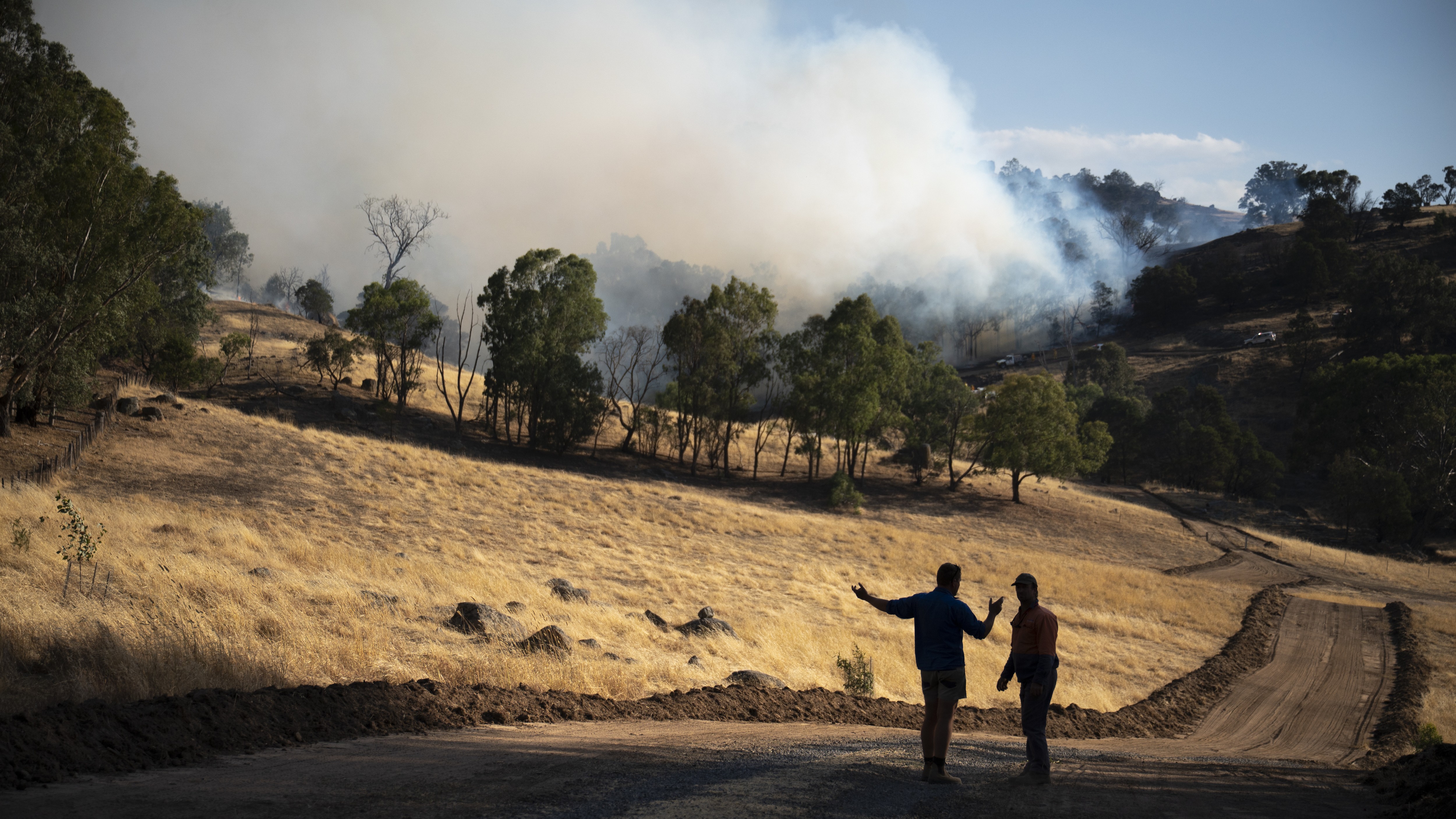 Two men stand on a road overlooking a burning property.