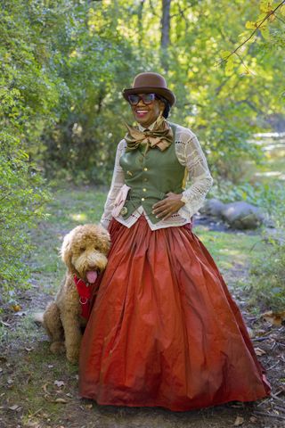 A photo of Arlinda McIntosh in an over 60 outfit featuring an orange ball skirt, white lace top, olive green waistcoat, a printed bow tie, and brown bowler hat.