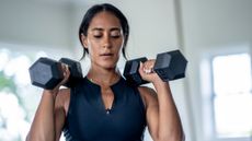 A woman in a sports vest holds a pair of heavy dumbbells at her shoulders. 