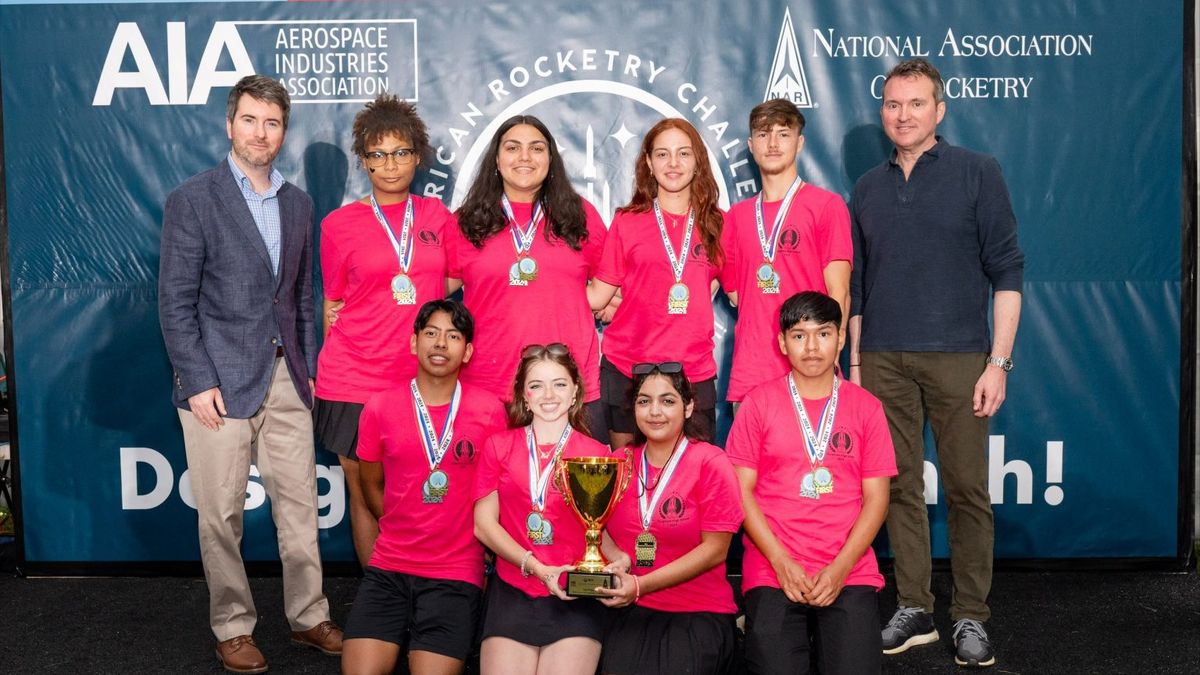 two men in dark tops stand on either side of students, all in pink, sorted in two rows of four, one standing behind, and the other kneeling in front. A banner behind them has the logo for the american rocketry challenge.