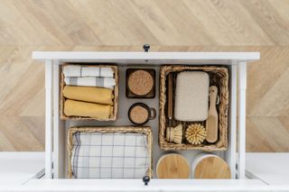 An open white, kitchen drawer organised with wicker baskets to help organise tea towels and scrubbing brushes.