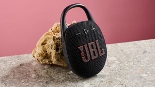 The JBL Clip 5 in black is pictured leaning against a sand coloured rock against a pink background,