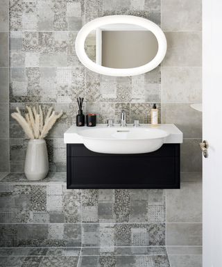 Gray tiled bathroom with unique mosaic effect, black and white sink, rounded white mirror and decorative vase with grasses