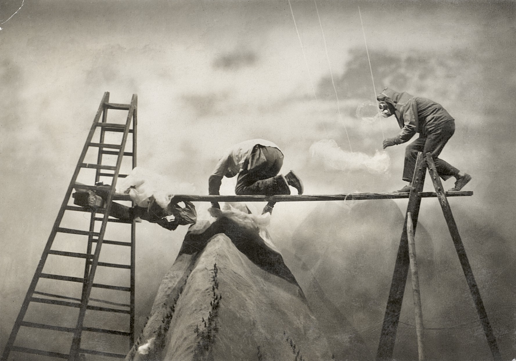 Two men balance on a wooden plank stretched between a ladder and a trestle above a model mountain peak, one wearing a hooded mask as smoke drifts around them against a painted cloudy backdrop.