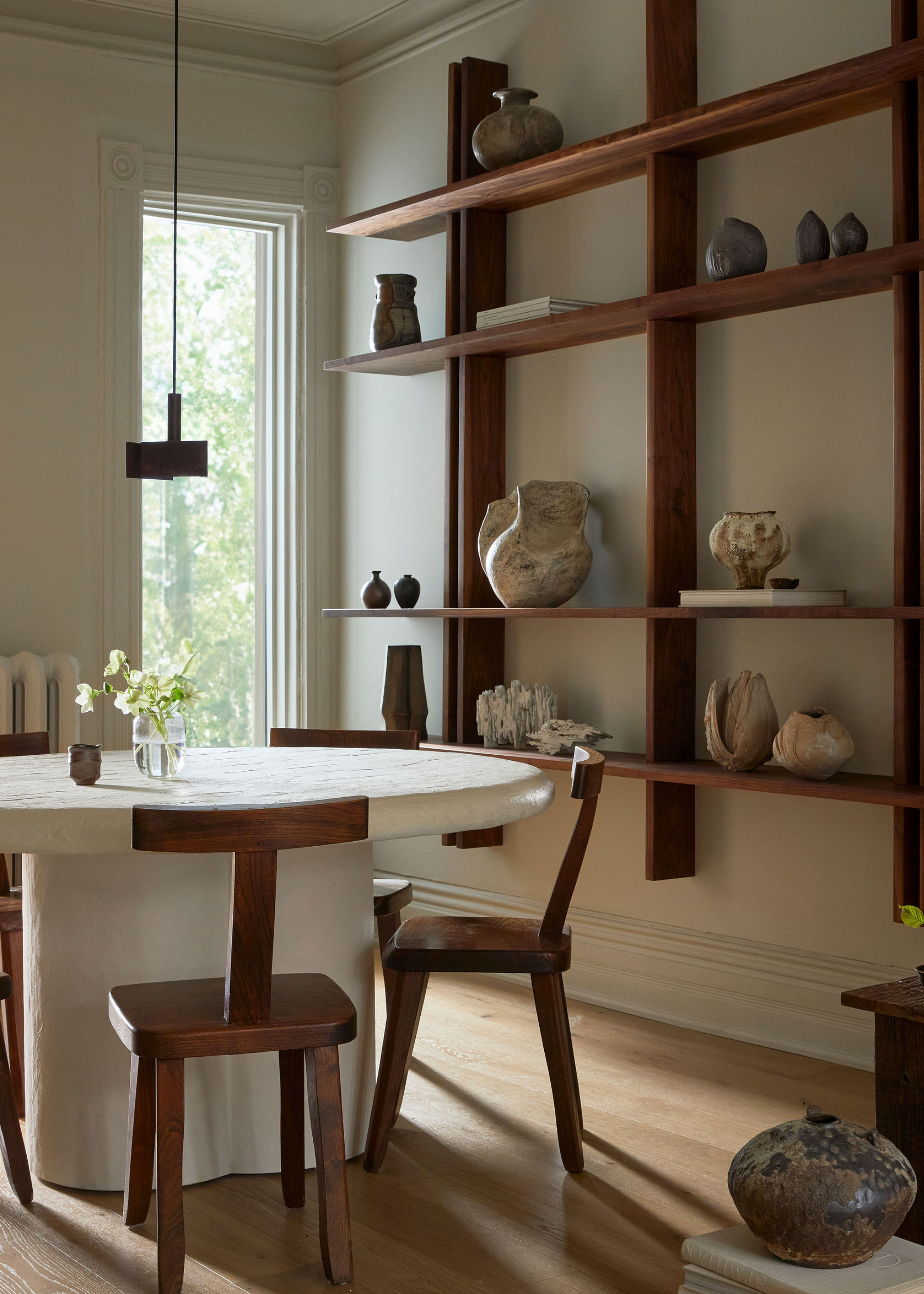 A dining room with a long window, a floating wood shelf of ceramic vases, and a white stone dining table with wood chairs