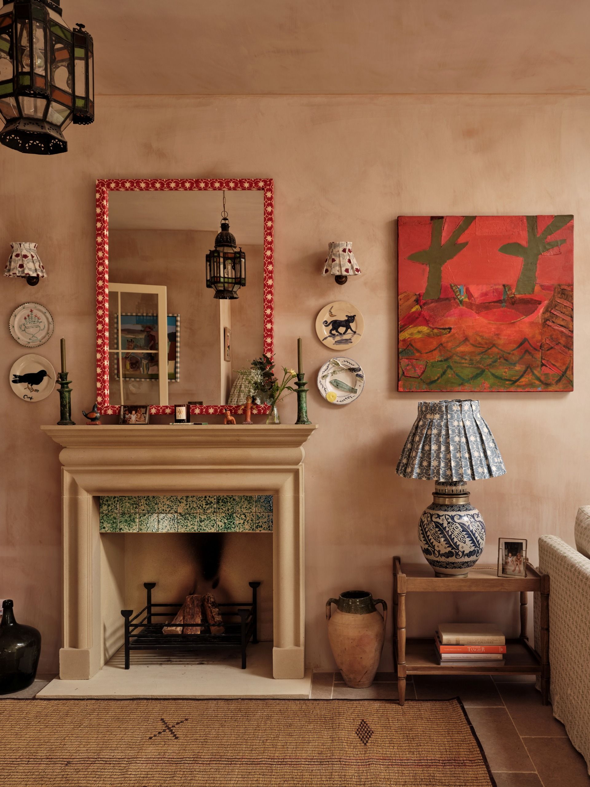 A period living room with pois, rattan, and checkered textile motifs, a fireplace decorated with a green and white tile frieze, lots of books, hung plates, and artwork, against argilla walls.