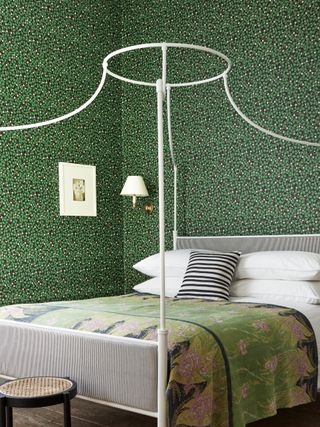 green bedroom with green ditsy wallpaper, white iron bed, green patterned bedspread, white artwork, white wall light