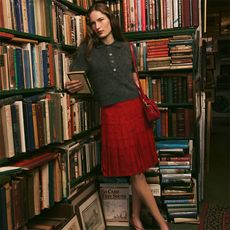 model wears gray brushed polo short-sleeve sweater and red pleated skirt while leaning against a book case. 