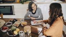 Two women sitting at a kitchen table. There is food on the table and one open laptop