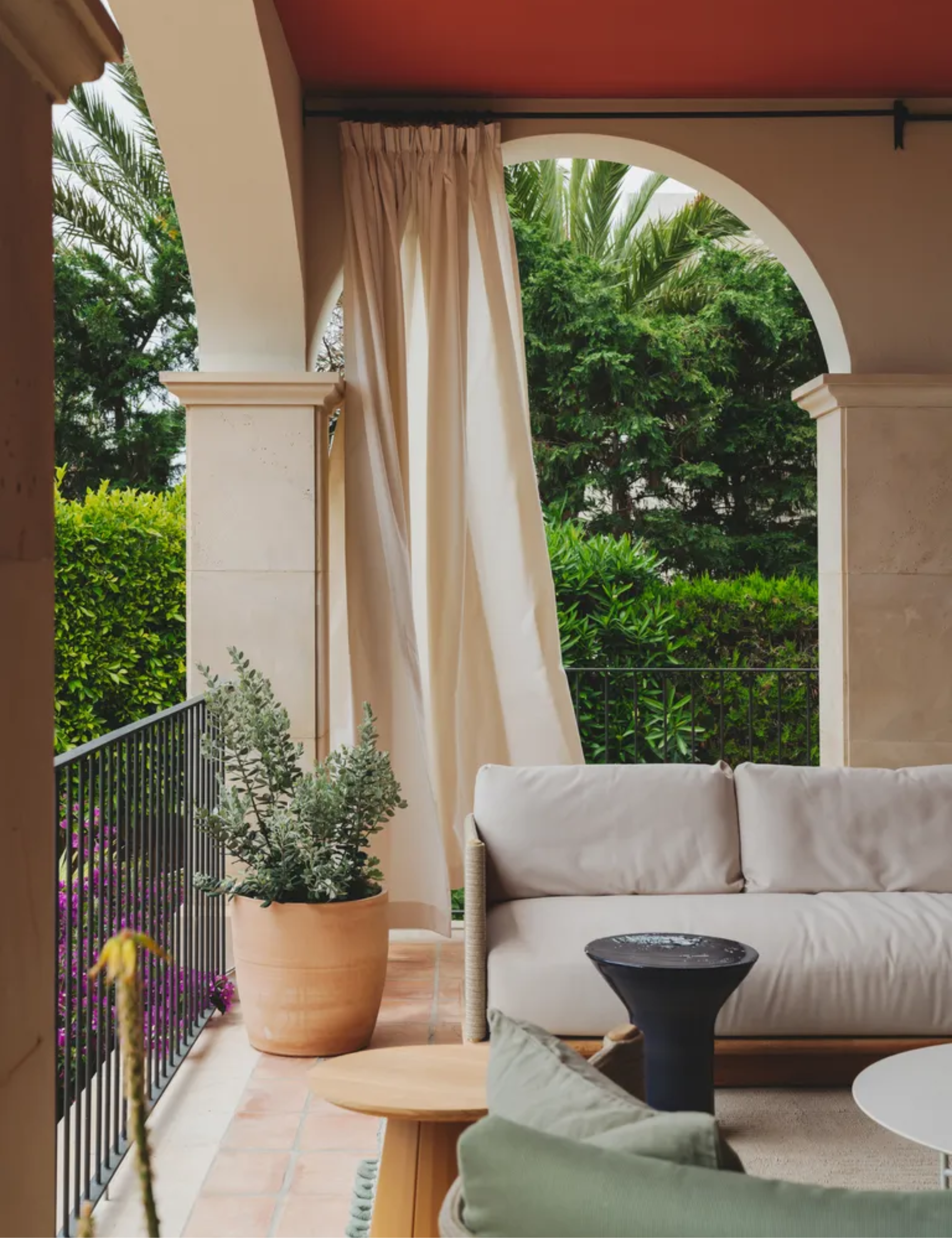 An outdoor terrace area with garden furniture, a ceiling painted red, and beige curtains hanging between the arches