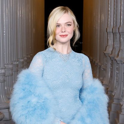Elle Fanning wearing a blue sheer dress and a matching fur shawl. Her hair is in an updo and two curls are framing each side of her face.