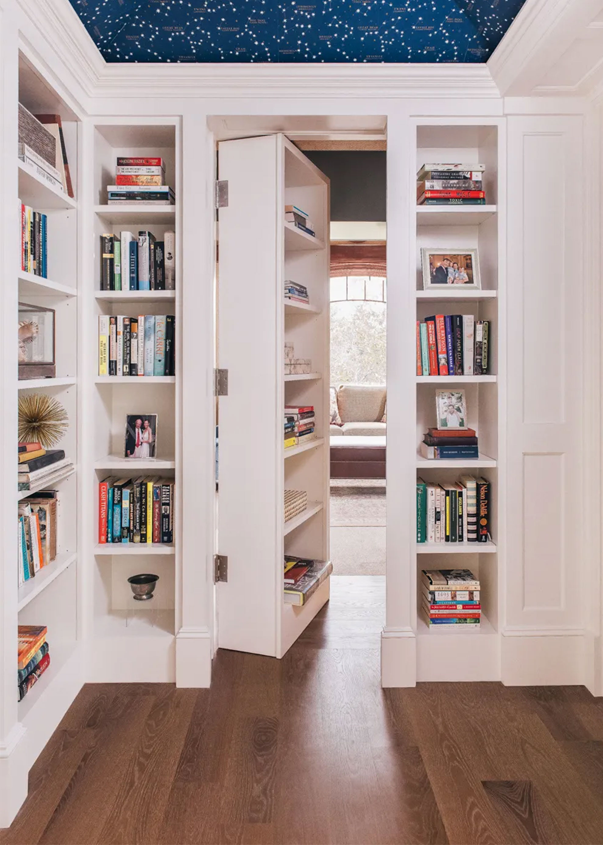 a bookcase door painted white filled with books blending in with the rest of the bookcases as a hidden door; the ceiling above is dark blue with speckled white and there is wooden flooring