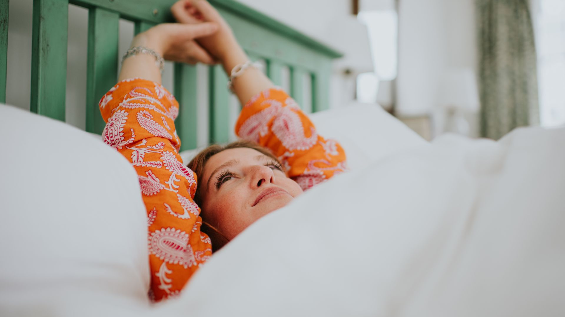 A woman lies in bed stretching her arms above her head after waking up in the morning.