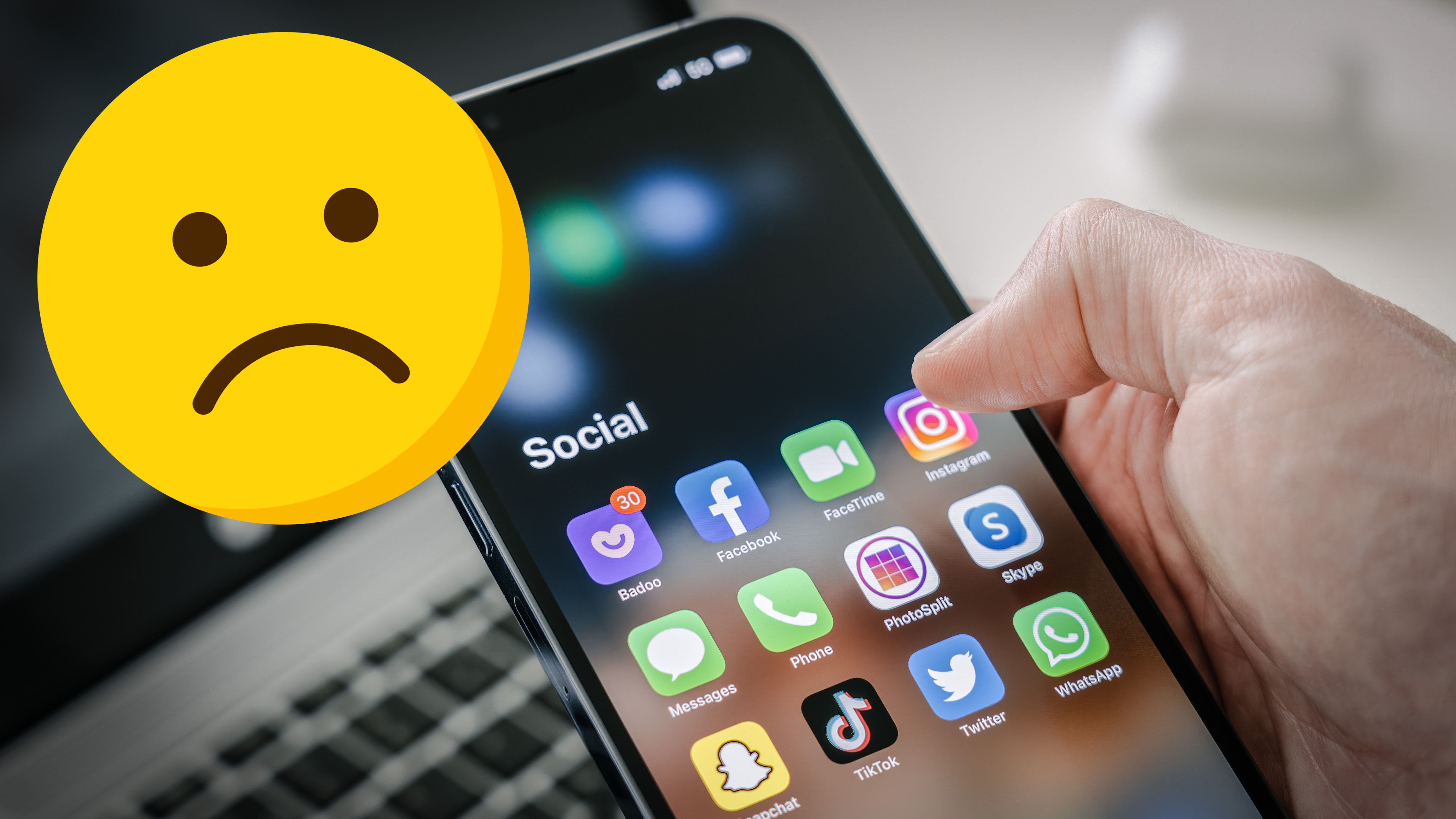 A man&#039;s hand holds a Iphone 13 Pro max with social media apps, against the background of a laptop. A sad face emoji has been superimposed over this image