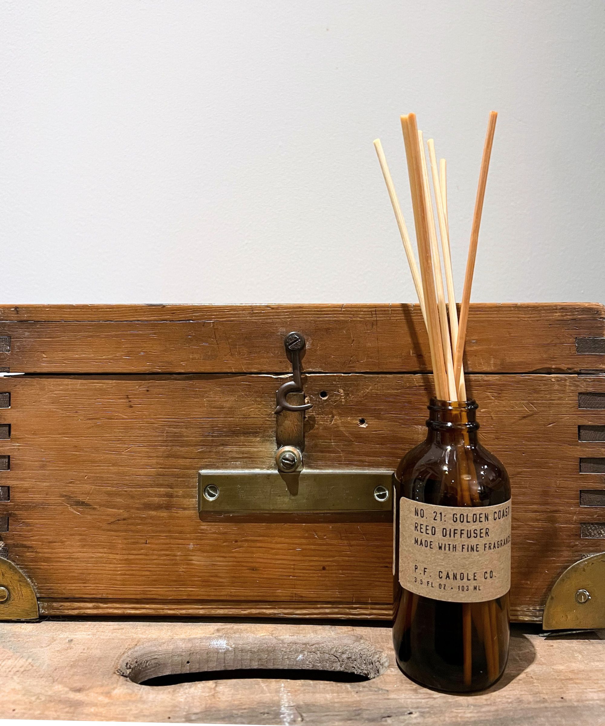 P.F. Candle Co. diffuser in brown glass bottle with brown reeds on a wooden box against a small wooden chest and grey wall.