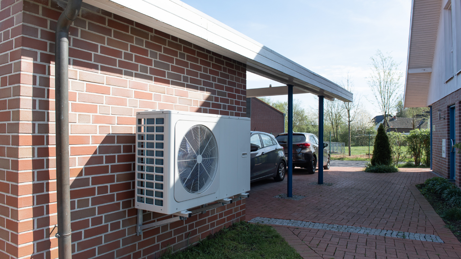 heat pump mounted on brick wall on side of house with carport, brick driveway and two cards in driveway