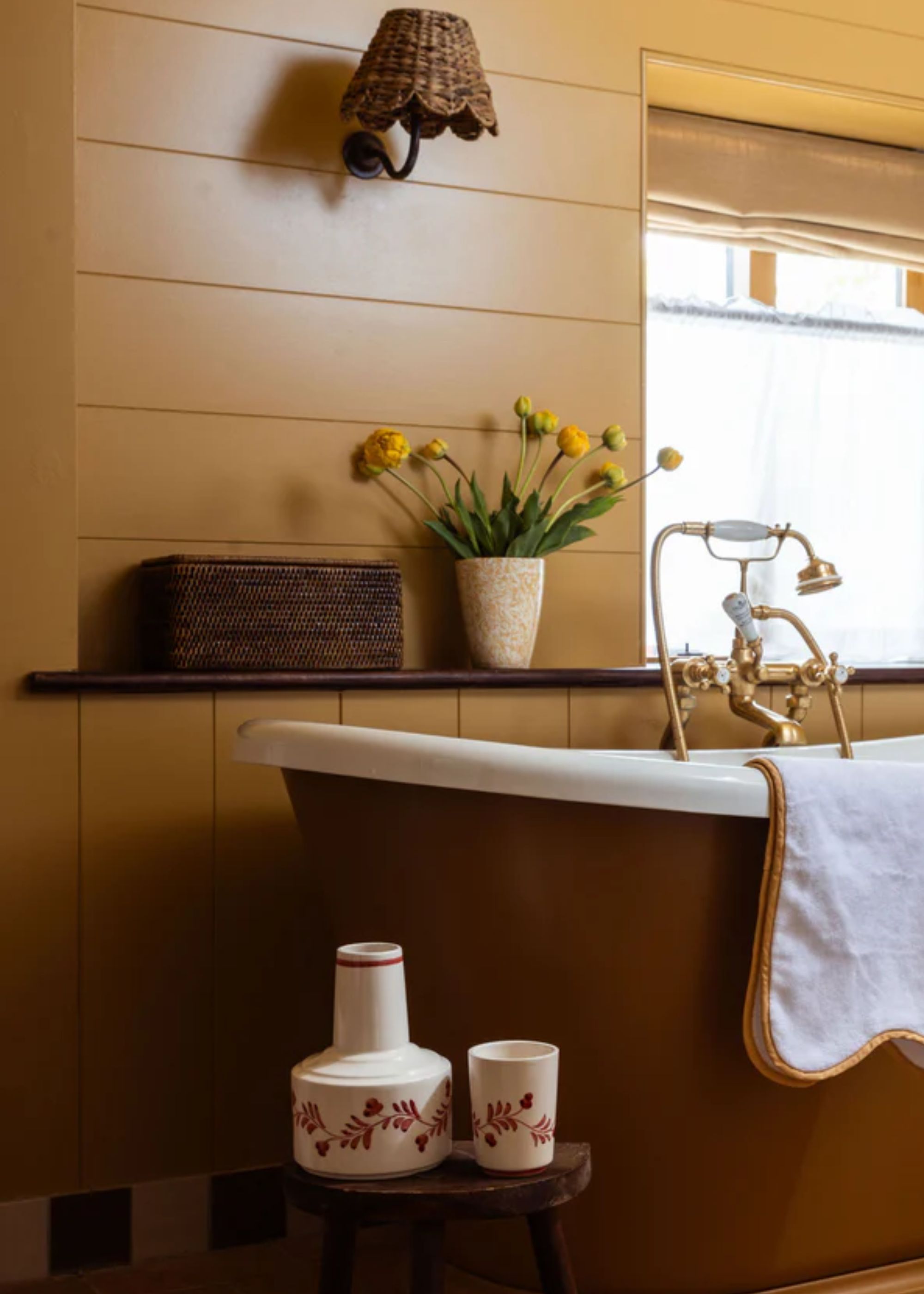 A bathroom with a bathtub and a basket placed on the shelving above it next to a pot of yellow flowers