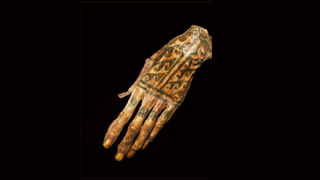 A disembodied mummified left hand has tattoos in black ink on the lower part of the fingers as well as on the back of the hand, in the shape of cresting waves