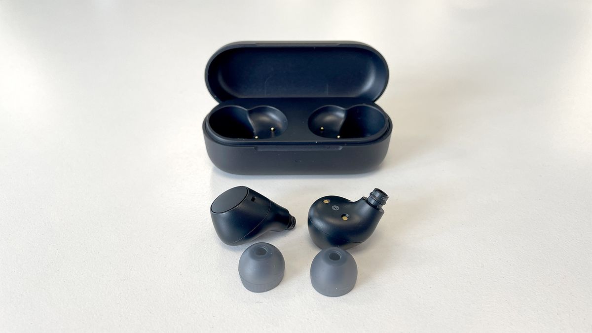 A black pair of Sony WF-C510 in-ear headphones out of the case with the ear tips removed. The case is in the background with the lid open. They are on a white background.