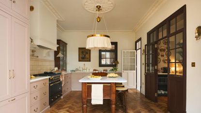 Beautiful cream kitchen with soft baby pink cabinets and range cooker, hardwood chevron floors, kitchen table island, crittal windows and doors through to dining room and statement crown molding and ceiling lamp