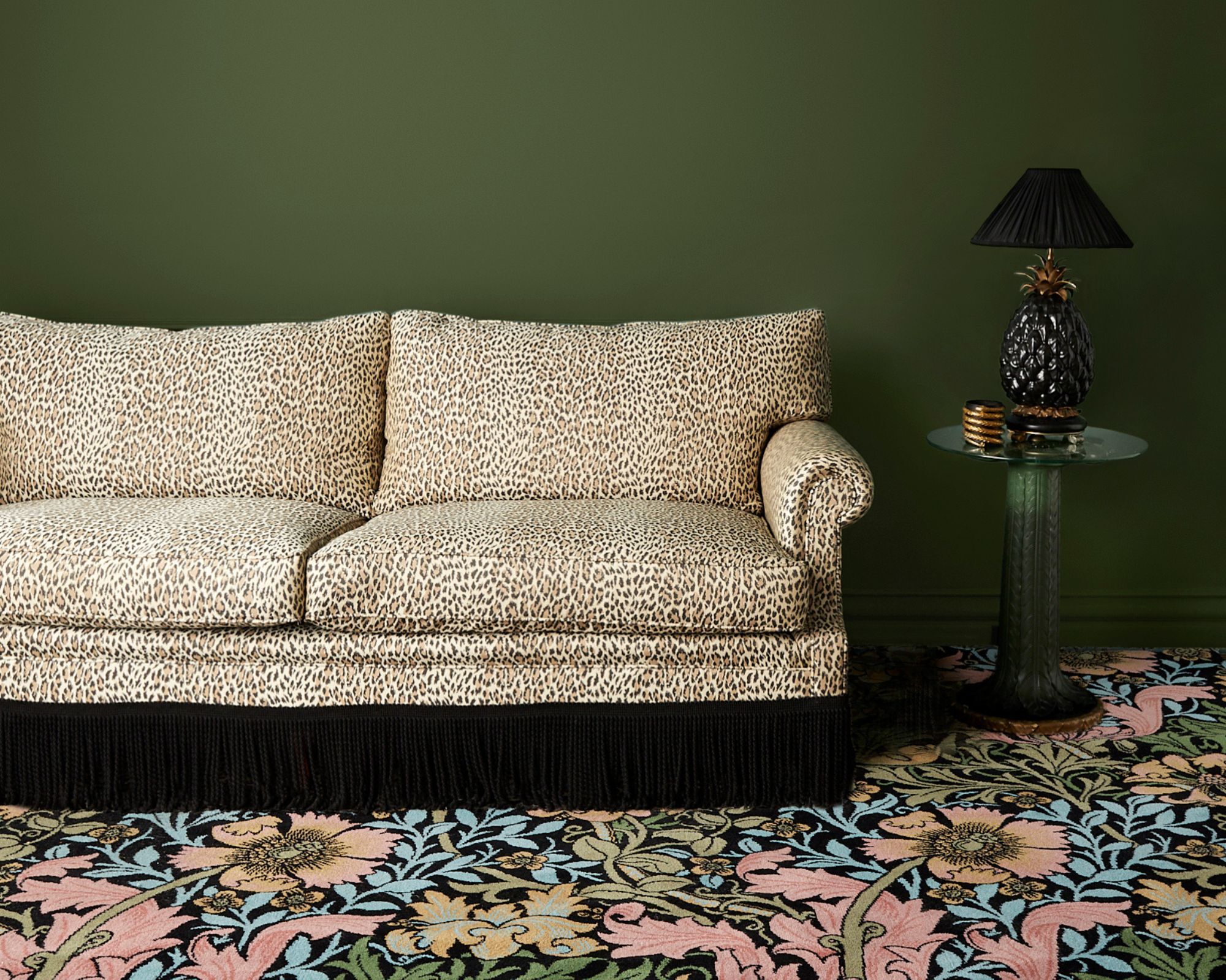 A leopard print sofa on colourful floral carpet, with a dark green painted wall and a side table with a black pineapple shaped lamp base with a black lampshade.