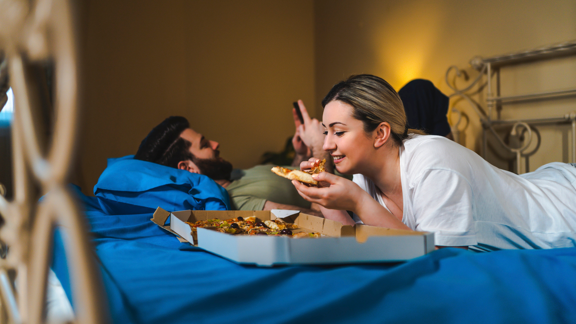 A woman lying on her front in bed next to a partner eating a pizza at night