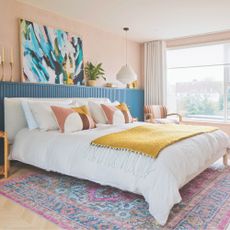 Bedroom with pink walls, blue panelling behind bed, colourful cushions and mustard throw. Artwork on wall and pendant lighting