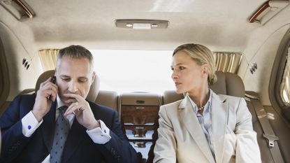 A high-net-worth couple look concerned in the back of a limo while he's on the phone.