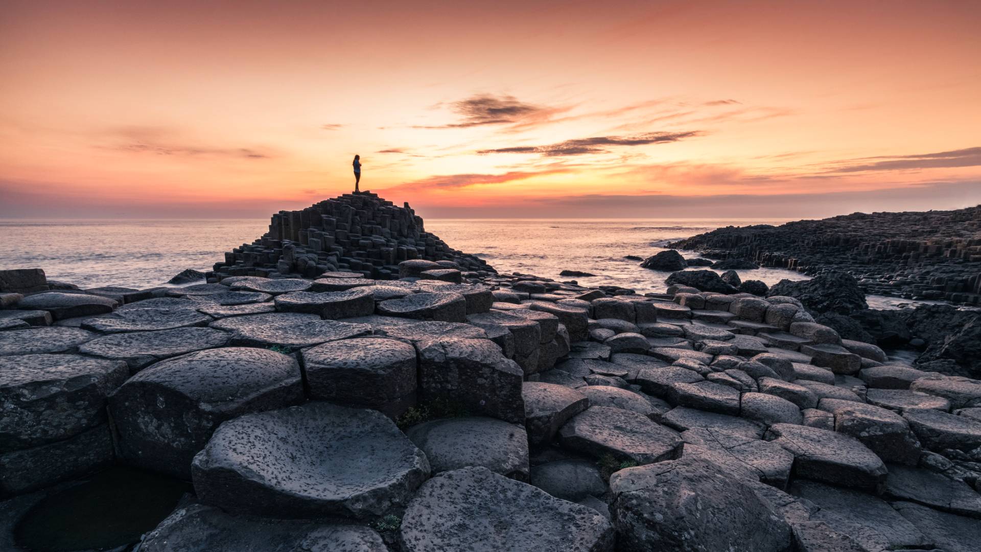 A person stands atop the hexagonal basalt columns of Giant&amp;rsquo;s Causeway in Northern Ireland at sunset, with a colorful sky and calm sea in the background.