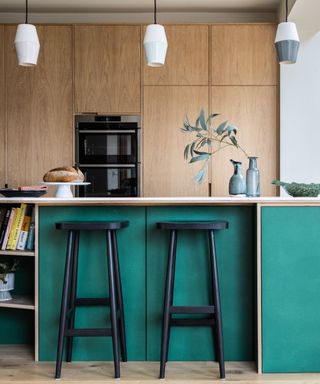 A kitchen with a dark green ktichen island with books on the shelves and two black stools, three white pendant lights, and light wooden cabinets behind it
