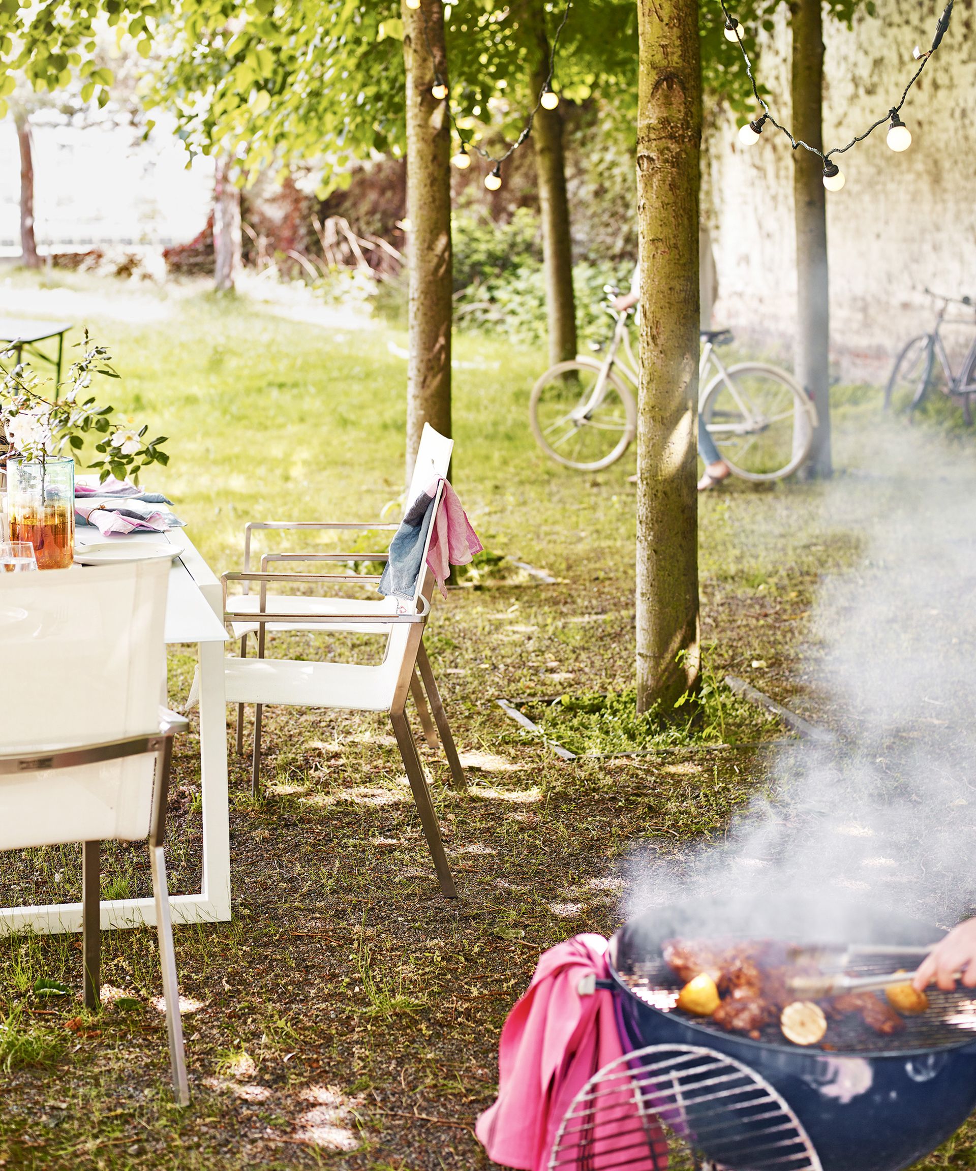 A lawn area with a white dining table, a barbecue and a line of trees