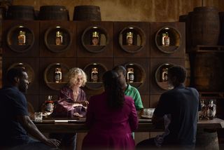 The tasting room of a well-appointed distillery. Everything is decorated in shades of teak, and there are ten different bottles of the distillery'srum on display. There is a large tasting table in the foreground. Francesca (Siobhán Redmond) and Cora (Madeline Appiah) are standing behind it, while Patrick (Ansu Kabia), Janelle (Michelle Asante) and Antony (Tienne Simon) are sitting on the other side, with their backs to the camera.