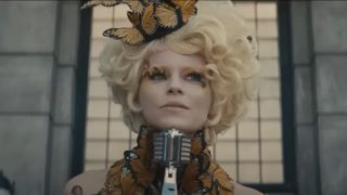 Elizabeth Banks stands in front of a microphone, in full Effie glam, in The Hunger Games: Catching Fire.